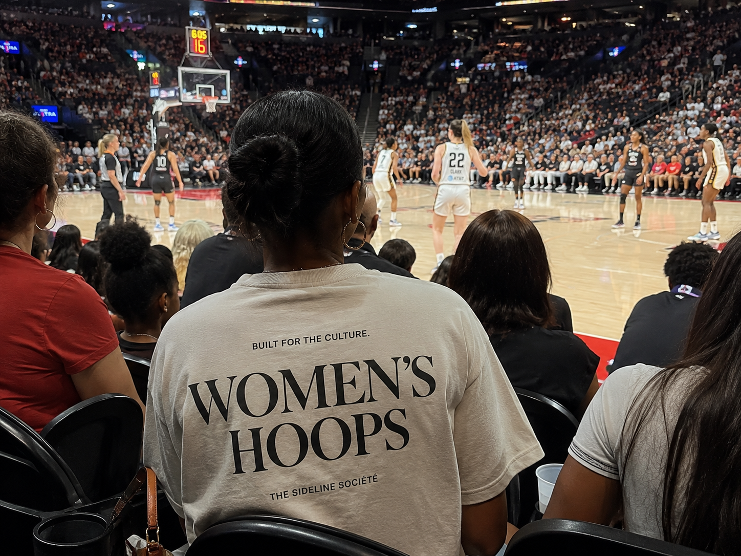 Woman wearing a cream Women’s Hoops T-shirt by The Sideline Société while watching a live women’s basketball game from courtside.